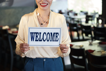 Crop view of happily smiling waitress in elegant clothes showing white Welcome sign in hands on blurred background 