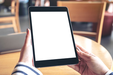 Mockup image of hands holding black tablet pc with blank white desktop screen on wooden table in cafe