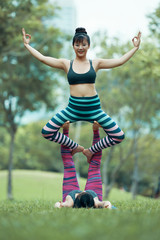 Beautiful young Asian woman sitting with stretched out arms on raised up legs of female lying on grass and looking at camera smiling on blurred background of park.