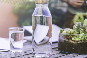 One bottle and glass of water on sunny wooden table outside on terrace close from flowers, cactus. Closeup