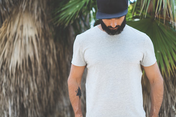 Bearded muscular hipster man model wearing gray blank t-shirt and a black baseball cap with space for your logo or design in casual urban style.Green palm and cactus garden on the background