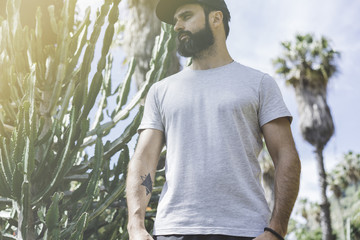 Bearded muscular hipster man model wearing gray blank t-shirt and a black baseball cap with space for your logo or design in casual urban style.Green palm and cactus garden on the background