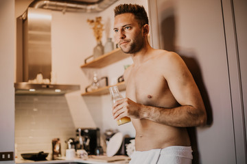 Attrctive young man with bare torso holding hand orange juice, looking out the window and smiling while standing in kitchen at home.