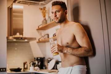 Sexy young man with bare torso holding hand orange juice, looking out the window and smiling while standing in kitchen at home.