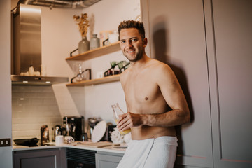 Shirtless hipster man drinking orange juice in the kitchen while leaning on the counter.