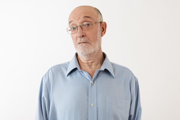 Picture of emotional elderly man with gray beard and bald head staring at camera with clueless facial expression, being at loss. Senior male in eyeglasses posing in studio, feeling confused