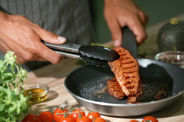 Man holding delicious roasted meat with tongs in kitchen, closeup