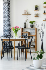 Industrial dining room interior with a table, black chairs, pink tulips, plants and shelves in the background