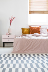 Bed with pink sheets and orange cushions standing in white bedroom interior with carpet, window...