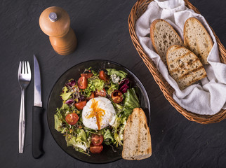  Green salad, ruccola, basil, spinach, poached egg, tomatos cherry and toast bread in round black plate, knife and fork near it on dark background. Healthy and tasty breakfast food.