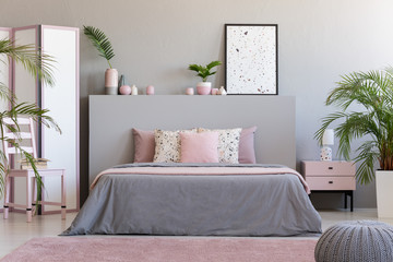 Poster on grey bedhead in bedroom interior with pink pillows on bed next to chair. Real photo