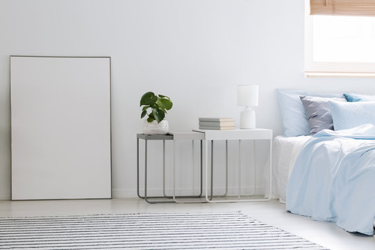 Empty mockup poster standing on the floor next to metal bedside table with fresh plant, books and lamp in white bedroom interior with carpet. Paste your graphic here