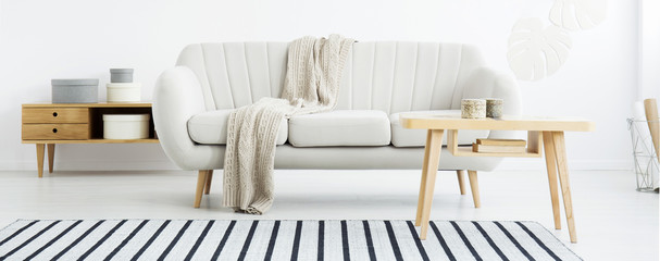 Real photo of living room interior with wooden cupboard with boxes, blanket on bright sofa, coffee table with books and striped carpet on the floor