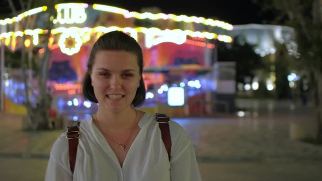 A close-up shot of a beautiful young woman smiling and walking in a shining amusement park in the evening with glowing carrousel on the background