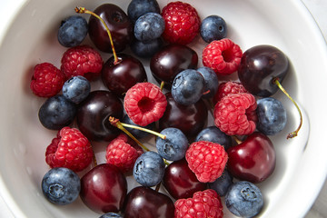 Close-up of ripe sweet fruits - cherry, raspberry, blueberries in the white plate on a gray stone background.
