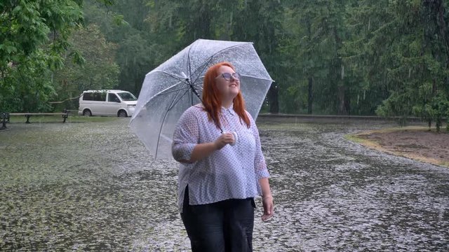 Fat ginger girl with glasses is walking in park under rain, watching up, holding umbrella