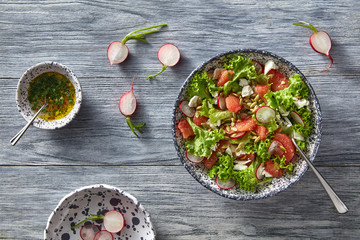 Fresh spring salad with radish, tomatoes, greens in ceramic bowl on gray wooden table. Flat lay.