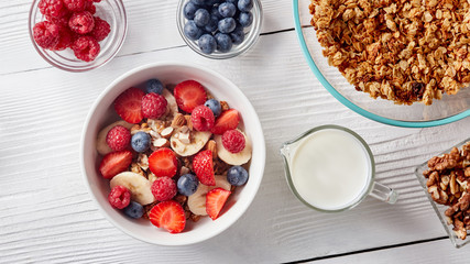 Homemade granola in a plate, sliced banana and strawberries, milk, almonds, blueberries - ingredients for natural breakfast on wthite table.