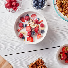 Ingredirnts for preparing healthy organic breakfast - granola, strawberries, honey, banana in white bowl with milk on white table. Flat lay.