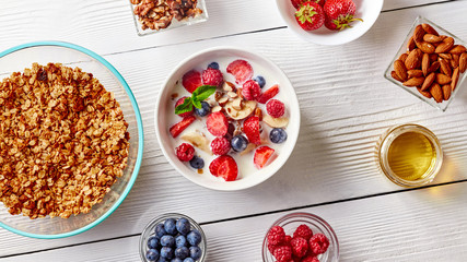 Cereal muesli and sliced bananas, strawberries, blueberries, raspberries, almonds and walnuts with milk in white ceramic bowl on white table. Top view.