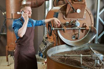 Portrait of handsome modern barista wearing apron speaking by phone while standing at coffee roasting machine in local roastery, copy space