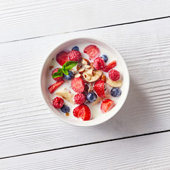 Colorful and tasty ready breakfast from corn muesli, berries, milk, almonds, slices banana on a bowl on a white wooden table with copy space. Flat lay.