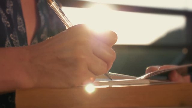 Woman's hand close-up, writing a note or letter to a notebook with a pen on a sunny day at sunset. A stack of scientific books, preparation for exams. Filling out the report