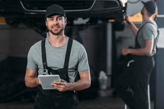 happy mechanic in overalls using digital tablet, while colleague working in workshop behind