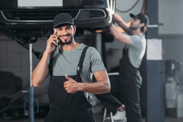 auto mechanic talking on smartphone and showing thumb up, while colleague working in workshop behind