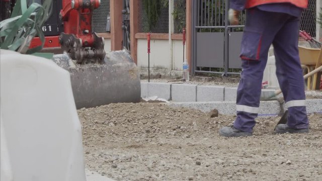 An excavator taking and dumping soil on a building site with a worker using a rake