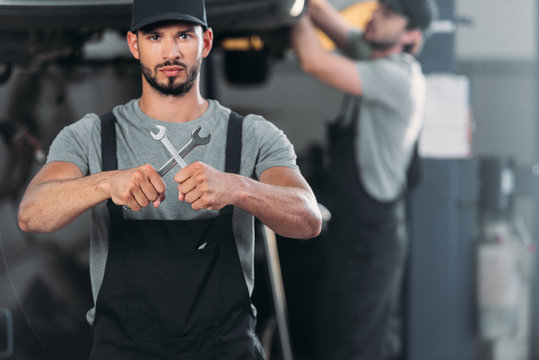 confident mechanic holding wrenches, while colleague working in workshop behind