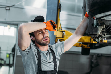 exhausted mechanic in uniform repairing car in workshop