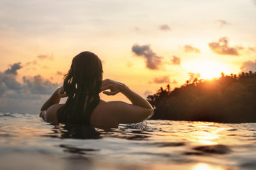 young woman from behind in indian ocean bathing and holding her hair during orange sunset with...