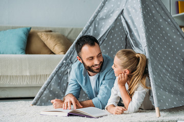 happy father and daughter lying on floor inside of teepee and reading book together at home © LIGHTFIELD STUDIOS