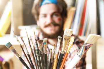 Dark-haired caucasian bearded hipster male artist in blue bandana hoilding many brushes in light...