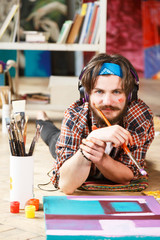 Caucasian dark-haired bearded hipster male artist in headphones and blue bandana lies on the floor...
