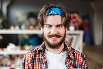 Portrait of handsome dark-haired bearded hipster male artist in blue bandana in his contemporary studio with many paintings