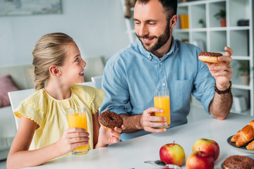 happy father and daughter eating doughnuts with orange juice at home