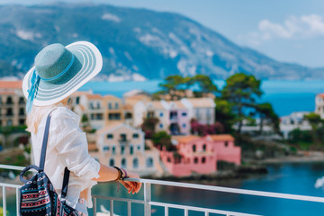 Tourist woman wearing blue sunhat and white clothes enjoying view of colorful tranquil village Assos on sunny day. Stylish female visiting Kefalonia in summer time on Greece travel vacation