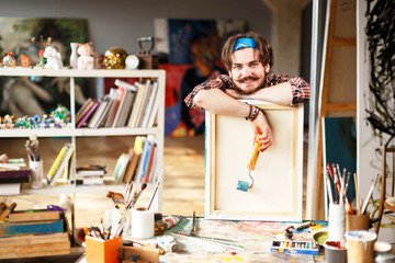 Dark-haired bearded hipster male artist in blue bandana holding dry brush and leane on canvas in...