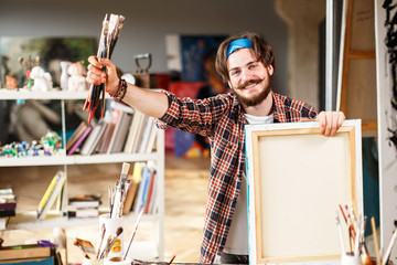 Handsome dark-haired bearded hipster male artist in blue bandana holding brush and smiling in his...