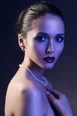 Beautiful brunette girl with naked shoulders and dark red lips make-up, wearing earrings and a pearl necklace, illuminated in blue.