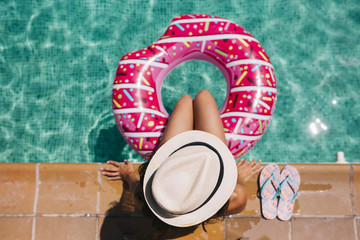top view of a woman relaxing in the pool with pink donuts in hot sunny day. Summer holiday idyllic. Enjoying suntan Woman in bikini and a hat. Holidays and summer lifestyle