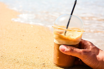A man's hand holds an iced coffee in a plastic glass with a black straw on a beach with yellow sand with a beautiful Ionian sea in the afternoon for enjoying and relaxing.