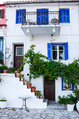 A white facade on old Greek houses with blue doors, windows in the afternoon delights tourists from all over the world. A staircase with various flowerpots and flowers.