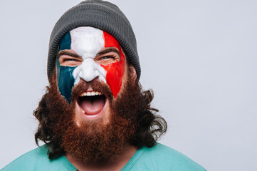 Face of young happy bearded man painted with flag of France. Football or soccer team fan, sport event, faceart and patriotism concept. Studio shot at gray background, copy space