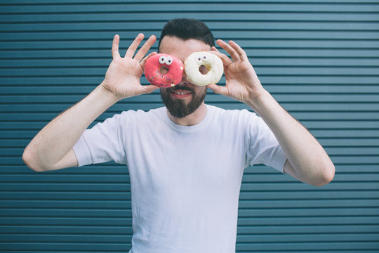 Funny picture of man holding two colorful donuts around his eyes. He is looking on camera and smiling. Guy has some fun. Isolated on striped and blue background.