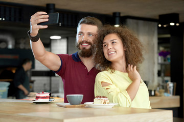 Front view of couple taking selfie while date in cafe. Drinking fragrant coffee, eating delicious cakes, looking positive, smiling. Boy wearing dark beard, girl having curly hair. Stylish interior.