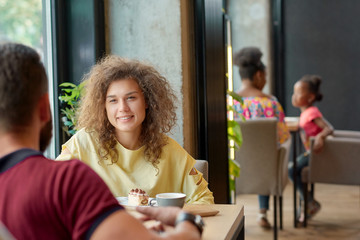 Curly girl sitting in cafe with boyfriend, drinking coffee, eating cakes. Model looking on boy sitting in front of her, wearing yellow blouse, smiling, feeling happy, satisfied, spending good time.