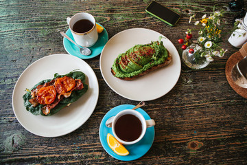 Sandwich with avocado and another sandwich with bacon and tomato next to two cups of tea on a wooden table